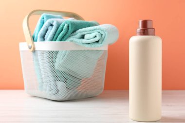 Çamaşır sepetinde havlular ve beyaz ahşap masada mercan arka planda bir şişe deterjan var.