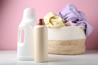 Çamaşır sepetinde elbiseler ve beyaz ahşap masada pembe arka planda bir şişe deterjan var.