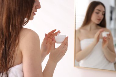 Havluya sarılı güzel bir kadın banyoda aynanın yanında kozmetik ürünler uyguluyor.