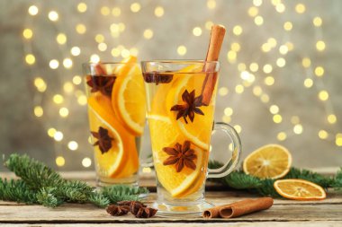 Portakallı, baharatlı ve köknar dallı lezzetli şaraplar ahşap masada gri arka planda, bulanık Noel ışıklarıyla, yakın plan. Bokeh etkisi