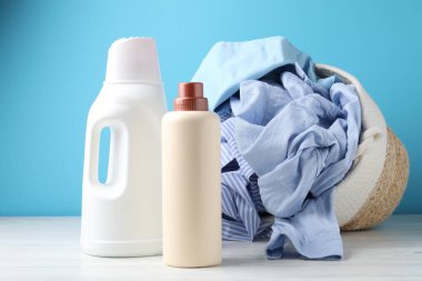 Çamaşır sepeti, beyaz ahşap masada açık mavi arka planda giysi ve şişe deterjan.