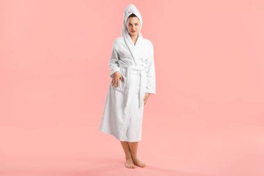 Pembe arka planda, saçı havluya sarılı bornoz giyen kadın.