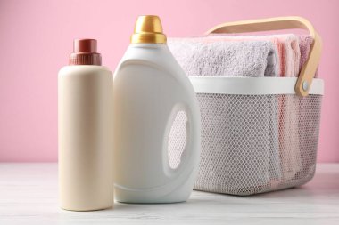 Çamaşır sepetinde havlular ve beyaz ahşap masada pembe arka planda duran deterjan şişeleri.