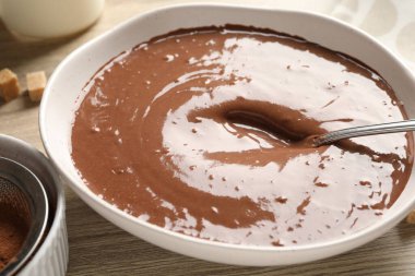 Liquid chocolate dough in bowl, cocoa powder and brown sugar on wooden table, closeup