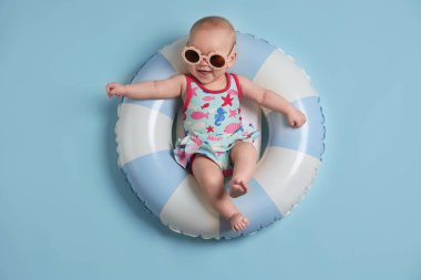 Şişirilebilir halkada güneş gözlüklü şirin bir bebek açık mavi arka planda, üst tarafta.