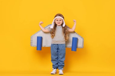 Kartondan uçak kanatları ve turuncu arka planda pilot olarak oynayan küçük bir kız.