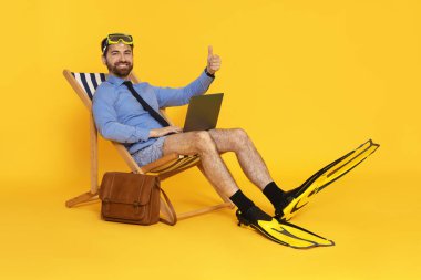 Dalış maskesi, dizüstü bilgisayarı ve yüzgeçleri turuncu arka planda güverte koltuğunda baş parmağını gösteren bir iş adamı.