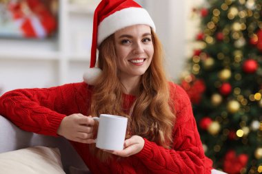Noel Baba şapkalı gülümseyen kadın koltukta sıcak bir bardak içecekle rahat bir odada bulanık ışıklara karşı. Mutlu Noeller ve mutlu yıllar.