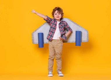 Karton kanatlı küçük çocuk turuncu arka planda pilotluk yapıyor.