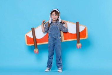 Karton kanatlı ve şapkalı küçük çocuk açık mavi arka planda pilotluk yapıyor.