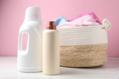 Çamaşır sepetinde elbiseler ve beyaz ahşap masada pembe arka planda bir şişe deterjan var.