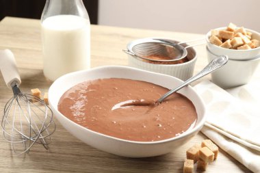 Kase içinde sıvı çikolata hamuru, süt, kakao tozu, esmer şeker ve çırpı, ahşap masada, yakın plan.