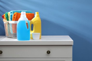 İçerideki mavi duvarın yanındaki şifonyerin üzerinde havlu ve şişe deterjan olan çamaşır sepeti.