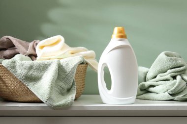 İçerideki yeşil duvarın yanındaki şifoniyerin üzerinde havlular ve bir şişe deterjan olan çamaşır sepeti.