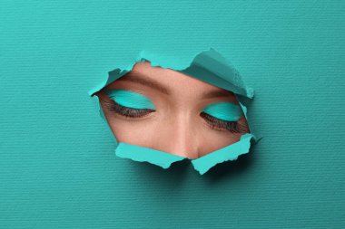 Parlak makyajlı genç bir kadın, yakın plan. Açık mavi kağıttaki yırtık delikten gözlerine bak.