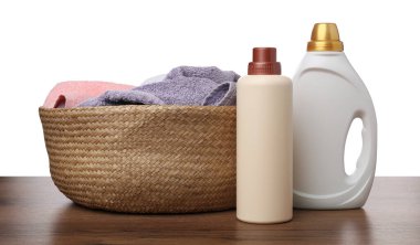 Çamaşır sepetinde havlular ve beyaz arka plandaki ahşap masada duran deterjan şişeleri.