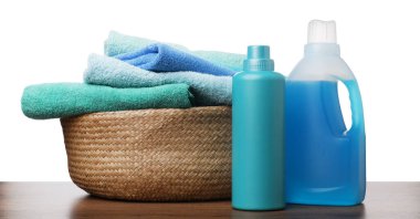 Çamaşır sepetinde havlular ve beyaz arka plandaki ahşap masada duran deterjan şişeleri.