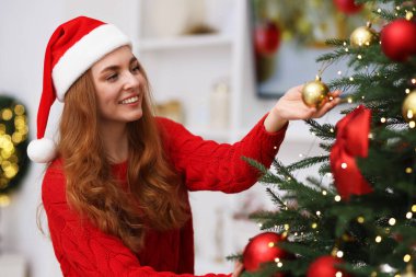 Noel Baba şapkalı gülümseyen kadın evdeki Noel ağacına güzel takılar takıyor.