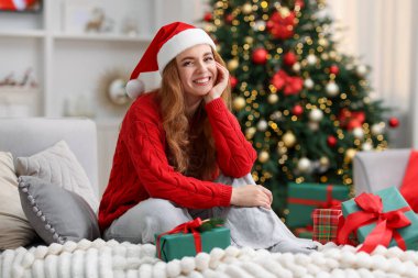 Noel Baba şapkalı gülümseyen kadın kanepede hediyelerle rahat bir odada bulanık ışıklara karşı.