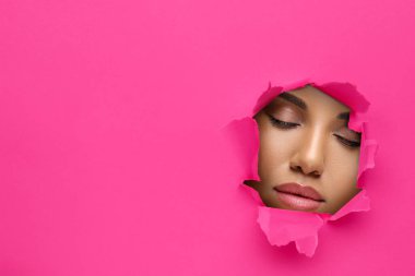 Güzel makyajlı genç bir kadın, pembe kağıttaki yırtık delikten bakıyor.