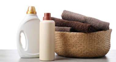 Çamaşır sepetinde havlular ve beyaz arka plandaki ahşap masada duran deterjan şişeleri.