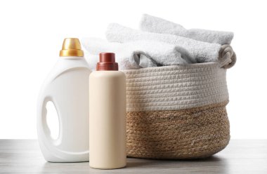 Çamaşır sepetinde havlular ve beyaz arka plandaki ahşap masada duran deterjan şişeleri.