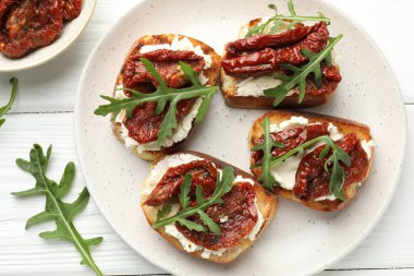 Krem peynirli nefis bruschettalar, güneşte kurutulmuş domatesler ve beyaz ahşap masada roka, düz uzanmış