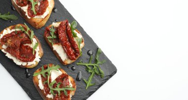 Krem peynirli nefis bruschettalar, güneşte kurutulmuş domatesler ve beyaz masa üzerinde roka. Metin için boşluk