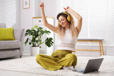 Evde, kulaklıklı, dans eden ve dizüstü bilgisayarlı mutlu bir kadın.