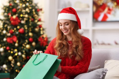 Noel Baba şapkalı gülümseyen kadın kanepenin üzerinde alışveriş torbasıyla rahat bir odada bulanık ışıklara karşı. Mutlu Noeller ve mutlu yıllar.