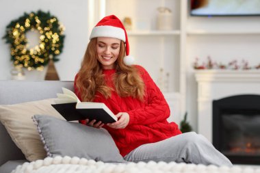 Noel Baba şapkalı gülümseyen kadın Noel için dekore edilmiş koltukta kitap okuyor.