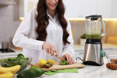 Gülümseyen kadın beyaz mermer masada smoothie için kivi kesiyor, blender içeride, yakın plan.