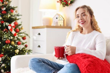 Koltukta bir fincan sıcak içecekle süveter giyen gülümseyen kadın Noel için dekore edilmiş odanın bulanık ışıklarına karşı. Metin için boşluk