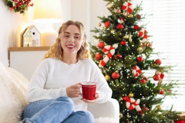 Koltukta bir fincan sıcak içecekle süveter giyen gülümseyen kadın Noel için dekore edilmiş odanın bulanık ışıklarına karşı. Metin için boşluk