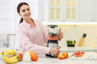 Gülümseyen kadın mutfaktaki beyaz mermer masada blender ile nefis smoothie yapıyor.