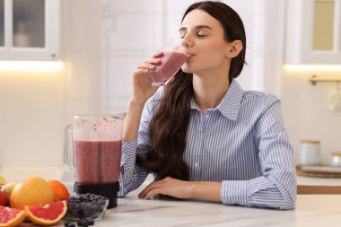 Beyaz mermer masada meyve suyu içen bir kadın. Mutfakta blender ve meyve var.