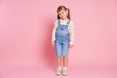 Cute little girl in jumpsuit standing on pink background