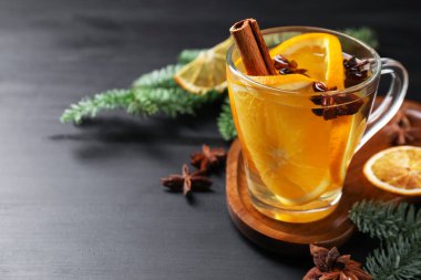 Delicious mulled white wine with slices of orange, spices and fir branches on black wooden table, closeup. Space for text