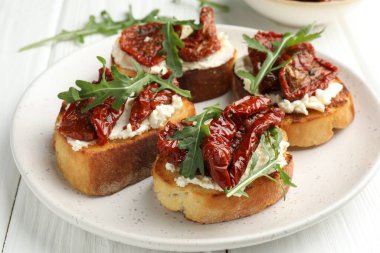 Krem peynirli lezzetli bruschettalar, güneşte kurutulmuş domatesler ve beyaz ahşap masada roka, yakın plan.