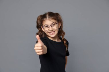 Gözlüklü gülümseyen kızın portresi gri arka planda baş parmağını gösteriyor