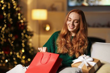 Kanepede Noel hediyeleri taşıyan süveterli bir kadın rahat bir odada bulanık ışıklara karşı. Metin için boşluk