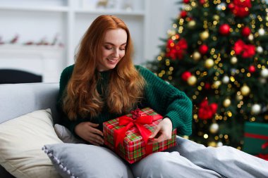 Kanepede Noel hediyesiyle gülümseyen kadın rahat bir odada bulanık ışıklara karşı.