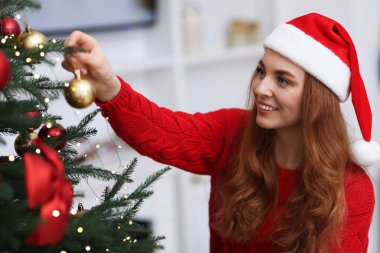 Noel Baba şapkalı gülümseyen kadın evdeki Noel ağacına güzel takılar takıyor.