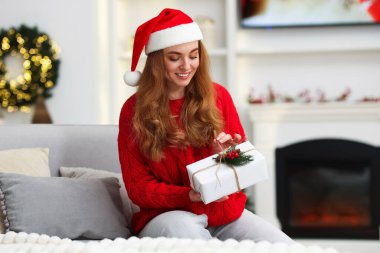 Noel Baba şapkalı gülümseyen kadın evdeki koltukta Noel hediyesiyle.