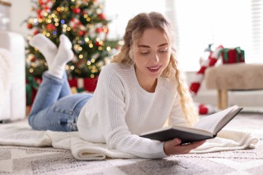 Süveter giyen, yerde kitap okuyan gülümseyen kadın Noel süslemeleri içinde bulanık ışıklara karşı.
