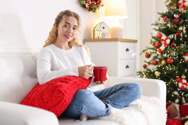 Kanepede bir fincan sıcak içecekle süveter giyen gülümseyen kadın Noel için dekore edilmiş odadaki bulanık ışıklara karşı.