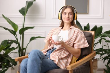 Elinde bir fincan aromatik kahve olan kadın evdeki koltukta müzik dinliyor.