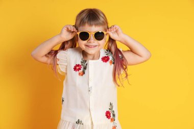 Gülümseyen küçük kız sarı arka planda güneş gözlüğü takıyor.