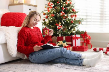 Süveter giymiş, elinde bir fincan sıcak içecek ile gülümseyen bir kadın yerde kitap okuyor Noel için dekore edilmiş odada bulanık ışıklara karşı.