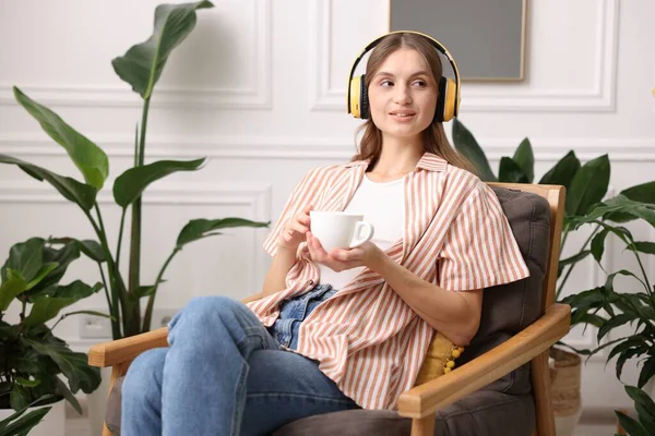 Elinde bir fincan aromatik kahve olan kadın evdeki koltukta müzik dinliyor.
